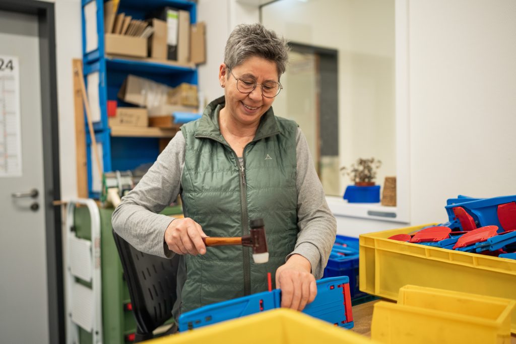 Mitarbeiterin mit einer mintgrünen Weste und Brille, die mit einem Hammer in einer Werkstatt steht