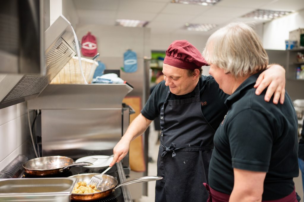 Zwei Mitarbeiter stehen in der Küche und bereiten Essen in einer Pfanne zu